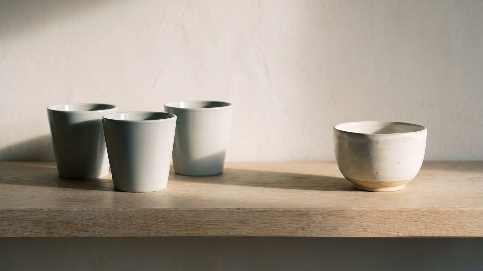 Three plain matching pale ceramic cups grouped together on a pale oak shelf, and standing apart from them a lower, wider hand-thrown stoneware bowl with a speckled glaze and an unglazed clay foot, against a lime-plaster wall in soft morning light.