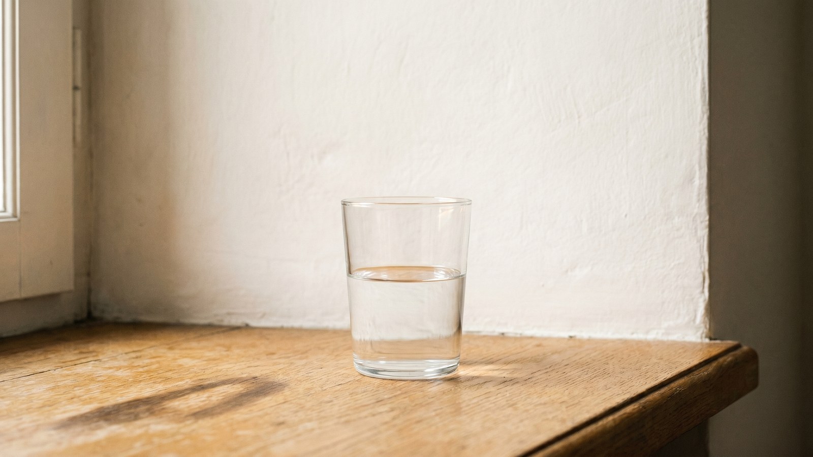 A plain clear-glass tumbler half-full of water on a warm French-oak windowsill in even morning light, a lime-plaster wall behind, one soft shadow across the sill.